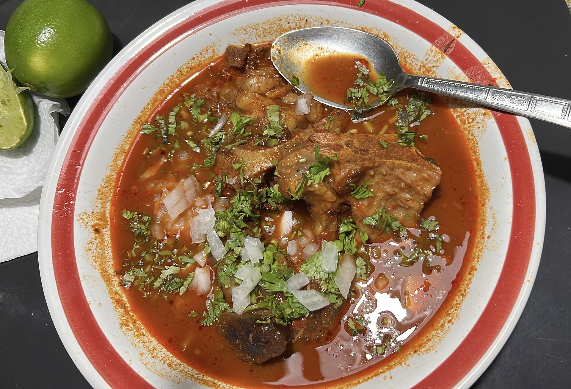 Bowl of Mexican birria stew in red chile broth topped with chopped onion and cilantro, with limes on the side